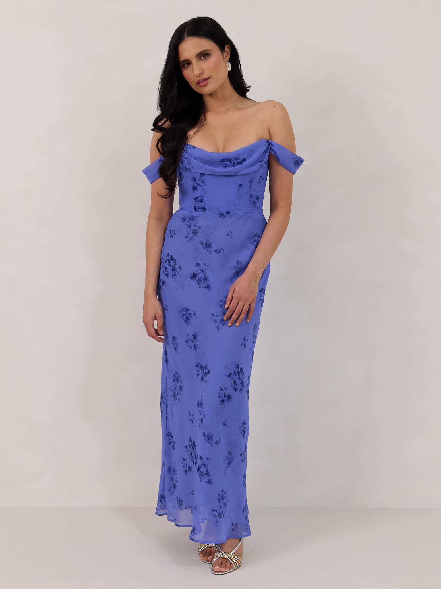 A woman standing against a white background wearing a periwinkle blue floral print dress. The dress has a scooped neckline with an off-the-shoulder strap, is fitted through the bodice and falls to ankle length. The fabric is chiffon. This is the Sutton Dress in Livia. 