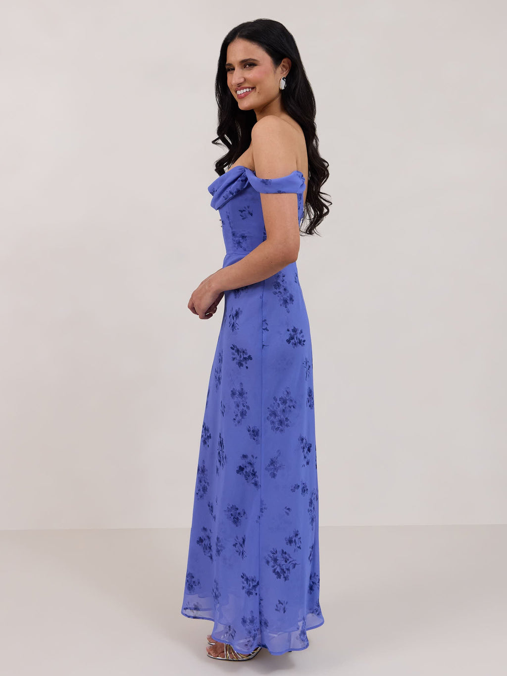 A woman standing facing the left against a white background wearing a periwinkle blue floral print dress. The dress has a scooped neckline with an off-the-shoulder strap, is fitted through the bodice and falls to ankle length. The fabric is chiffon. This is the Sutton Dress in Livia. 
