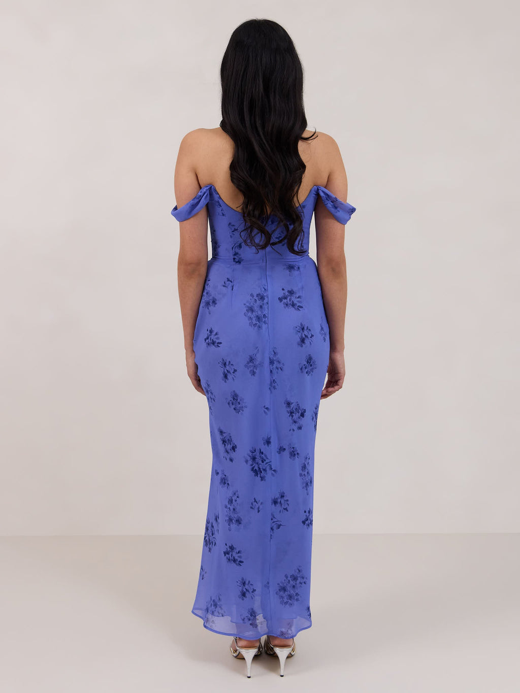 A woman standing facing the back against a white background wearing a periwinkle blue floral print dress. The dress has a scooped neckline with an off-the-shoulder strap, is fitted through the bodice and falls to ankle length. The fabric is chiffon. This is the Sutton Dress in Livia. 