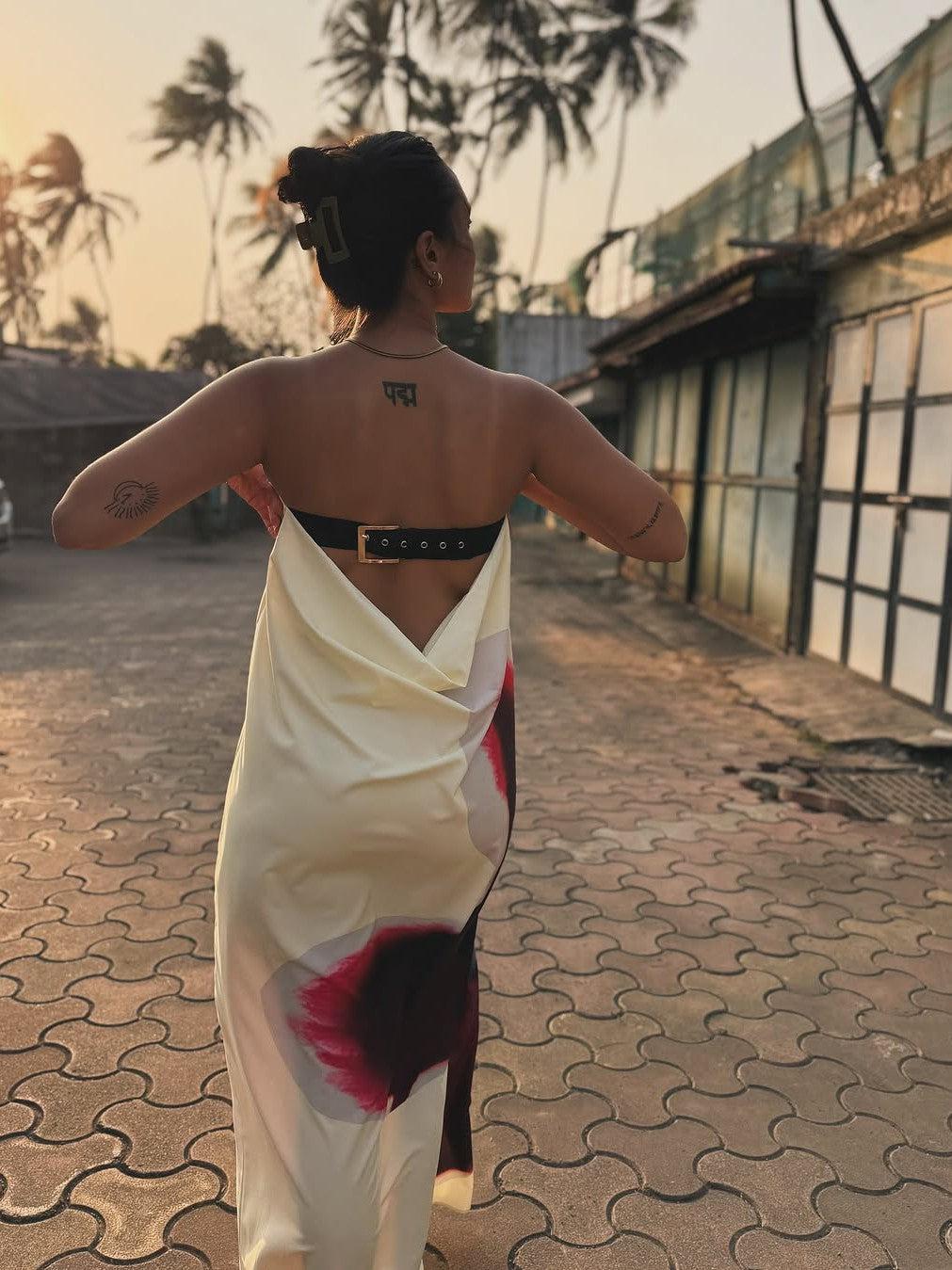 Back view of a woman in a cream floral print backless strapless maxi dress with a black belt, sunset palm trees.