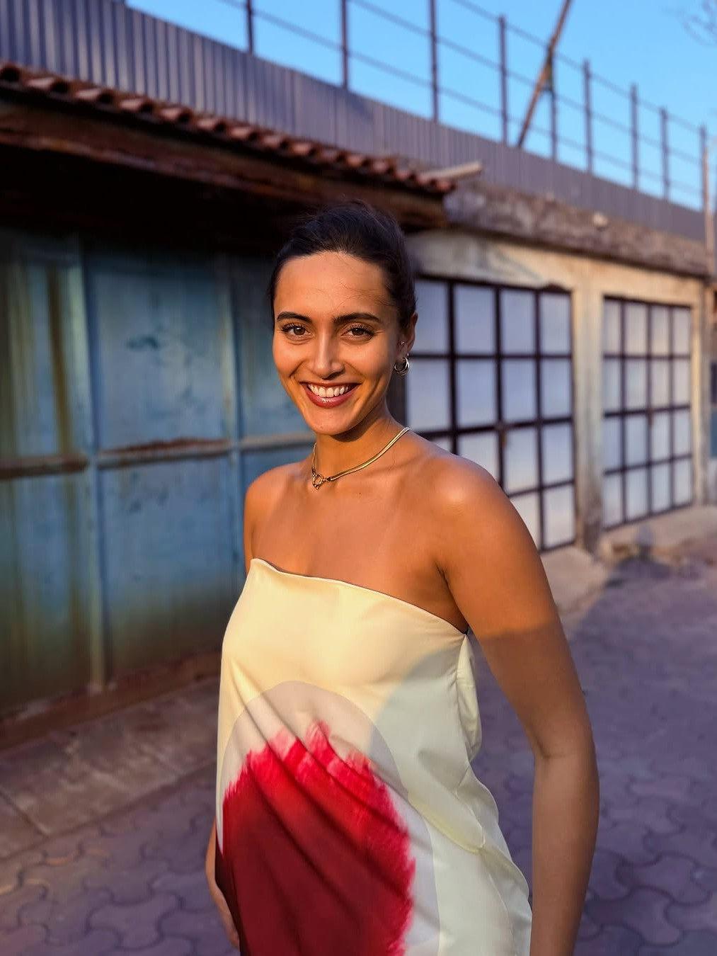 Smiling woman wearing a pale yellow strapless tube maxi dress with a bold red circular print, standing outdoors beside a grid-windowed building.