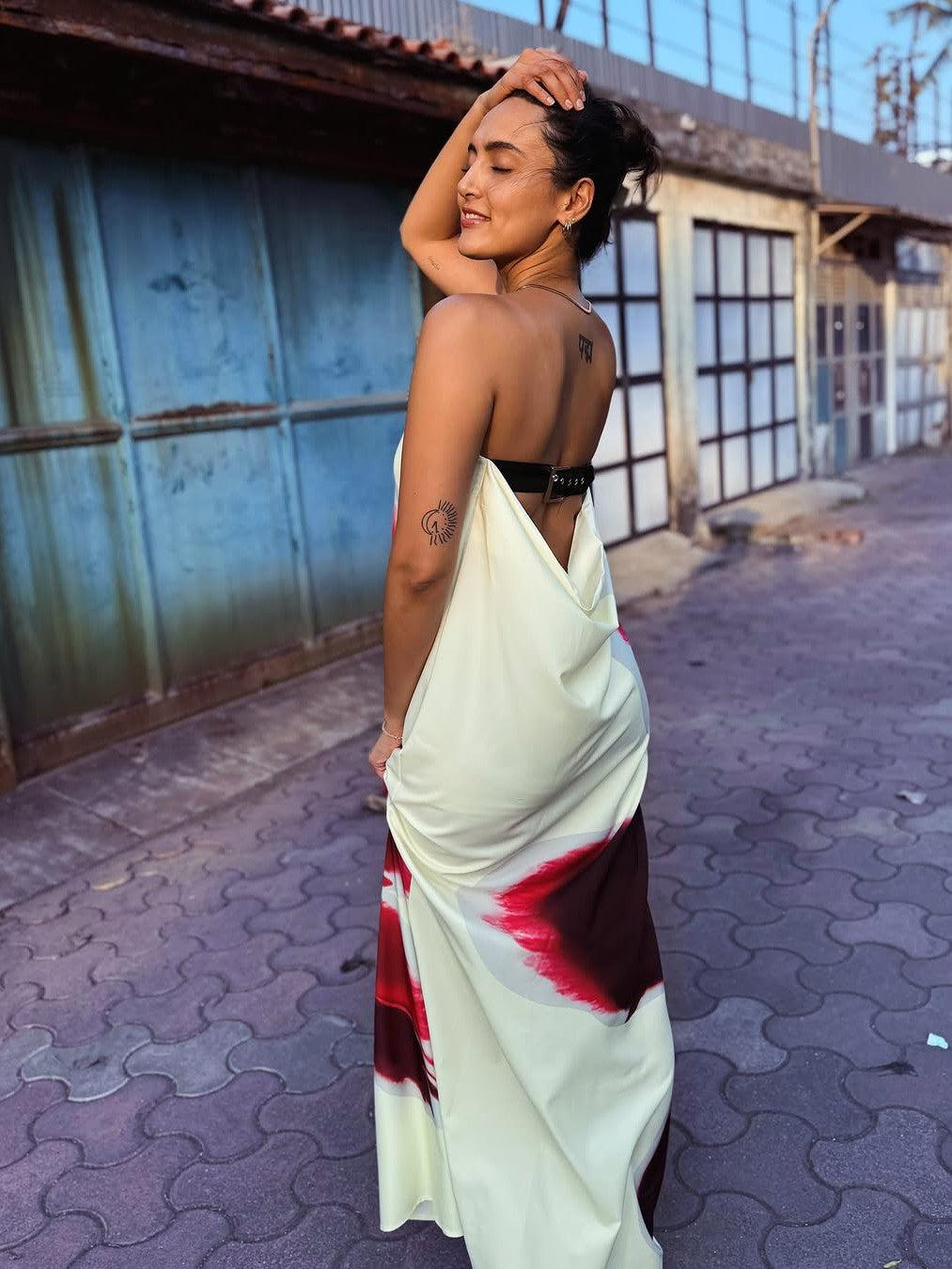 Woman in pale yellow backless maxi dress with floral print and black belt, standing in an urban alley.