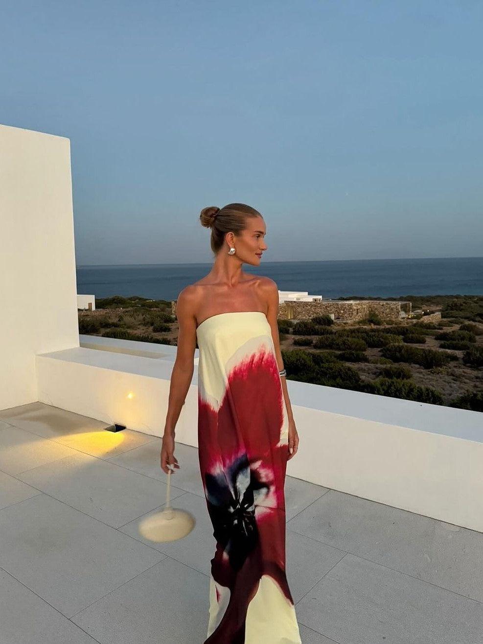 Woman in strapless backless tube maxi dress with cream, red and navy tie-dye floral print, standing on a white terrace by the sea, holding a bag.