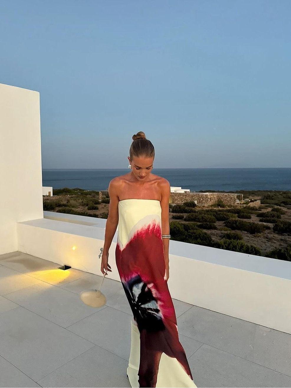 Cream strapless backless maxi dress with bold red and black floral print, worn by a model on a sea view terrace.