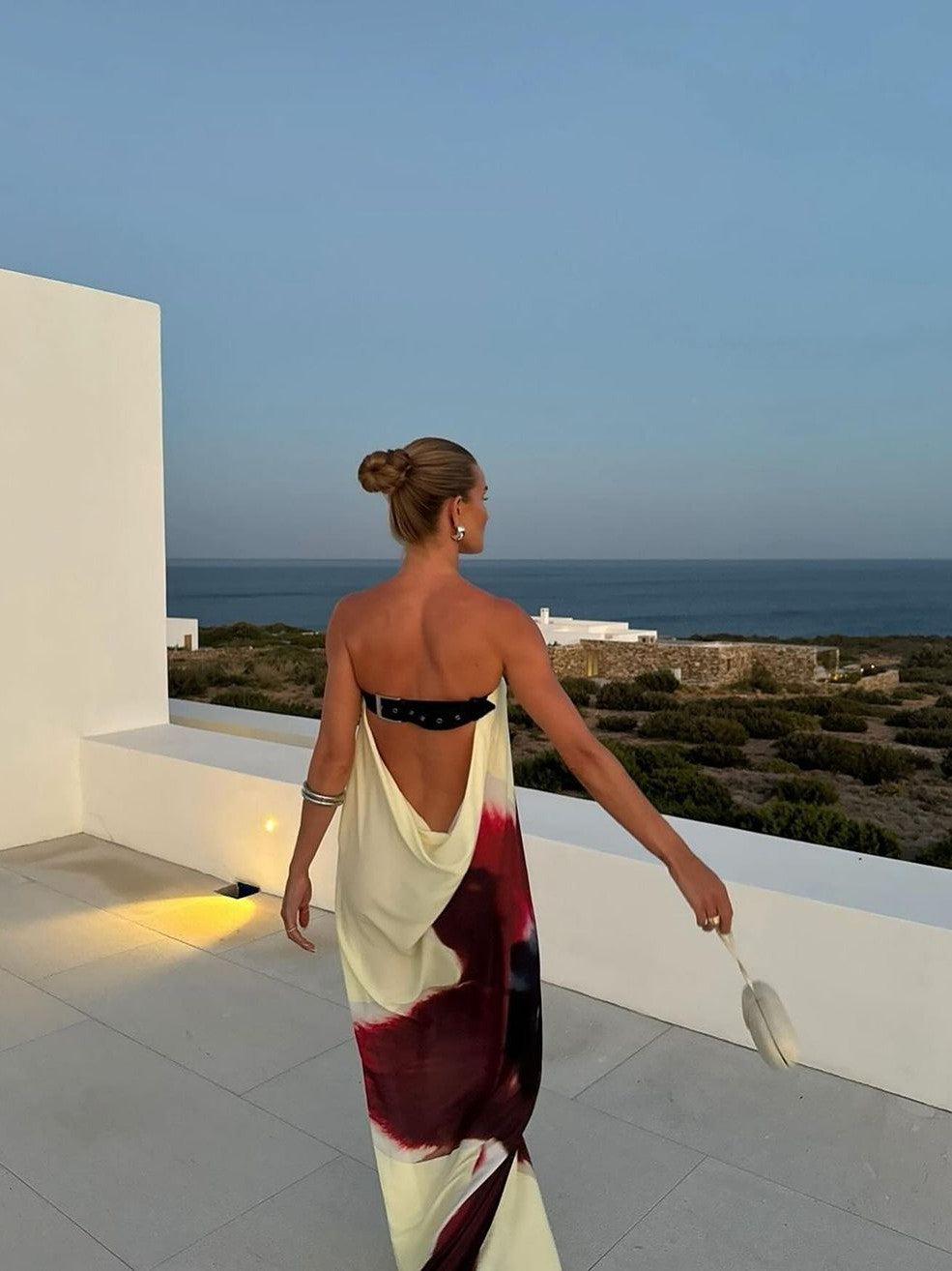 Backless draped floral print maxi dress in pale yellow with burgundy accents, paired with a black bandeau; ocean-view terrace backdrop.