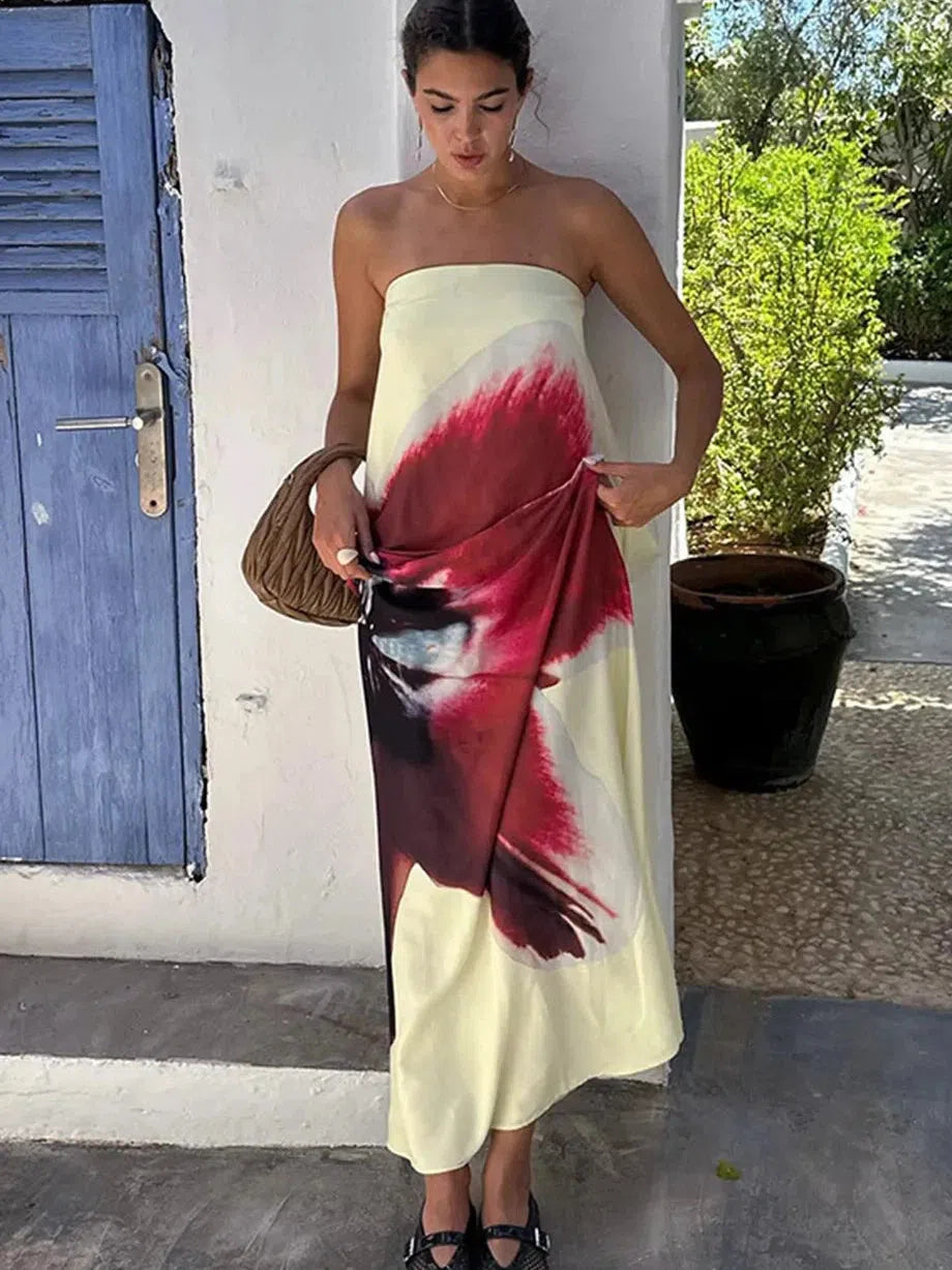 Strapless pale-yellow maxi dress with large red-pink abstract floral print, model holding a brown woven bag outdoors.